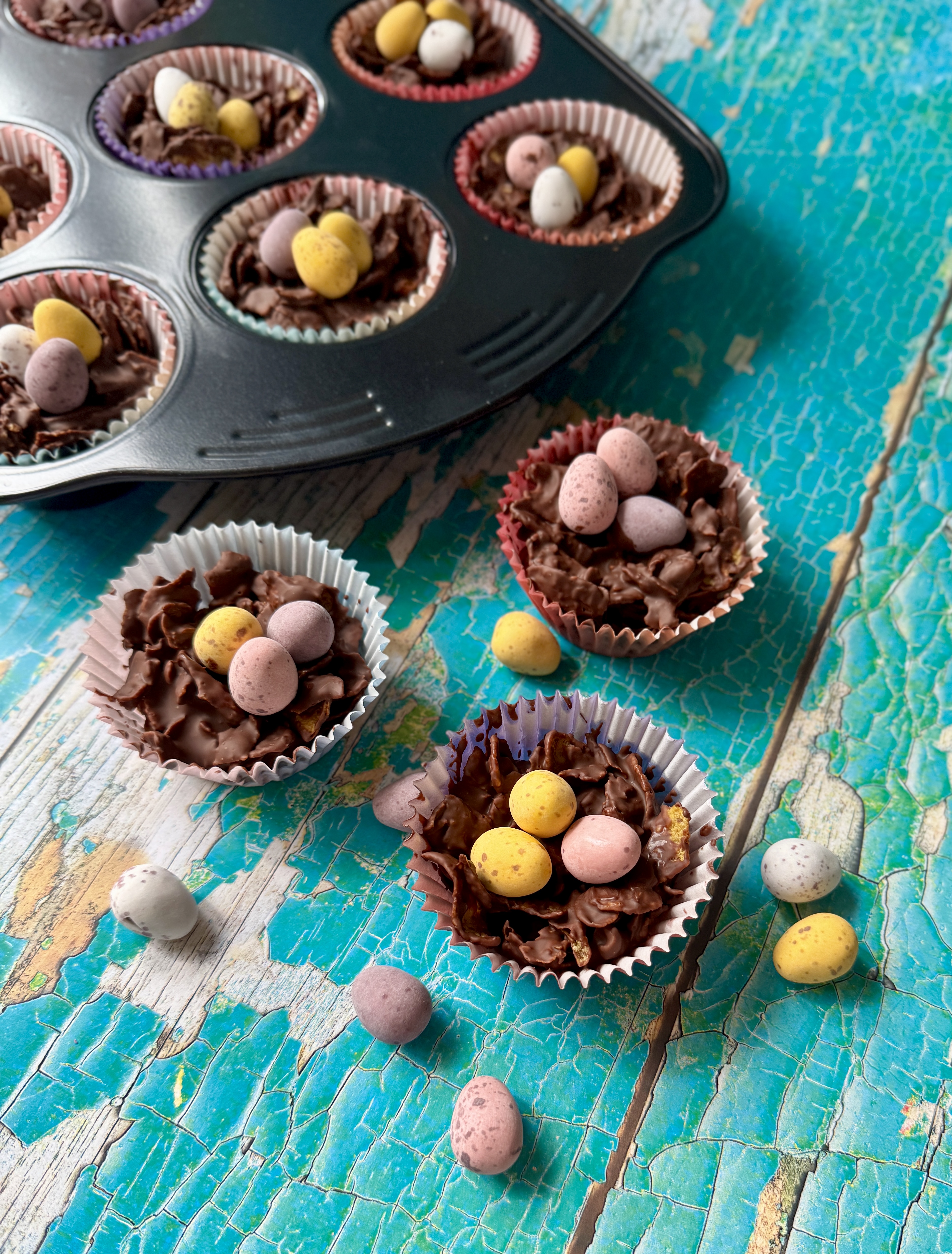Homemade Easter cornflake cakes in paper cases, topped with pastel Mini Eggs, with more cakes in a cupcake tray on a distressed turquoise wooden surface.